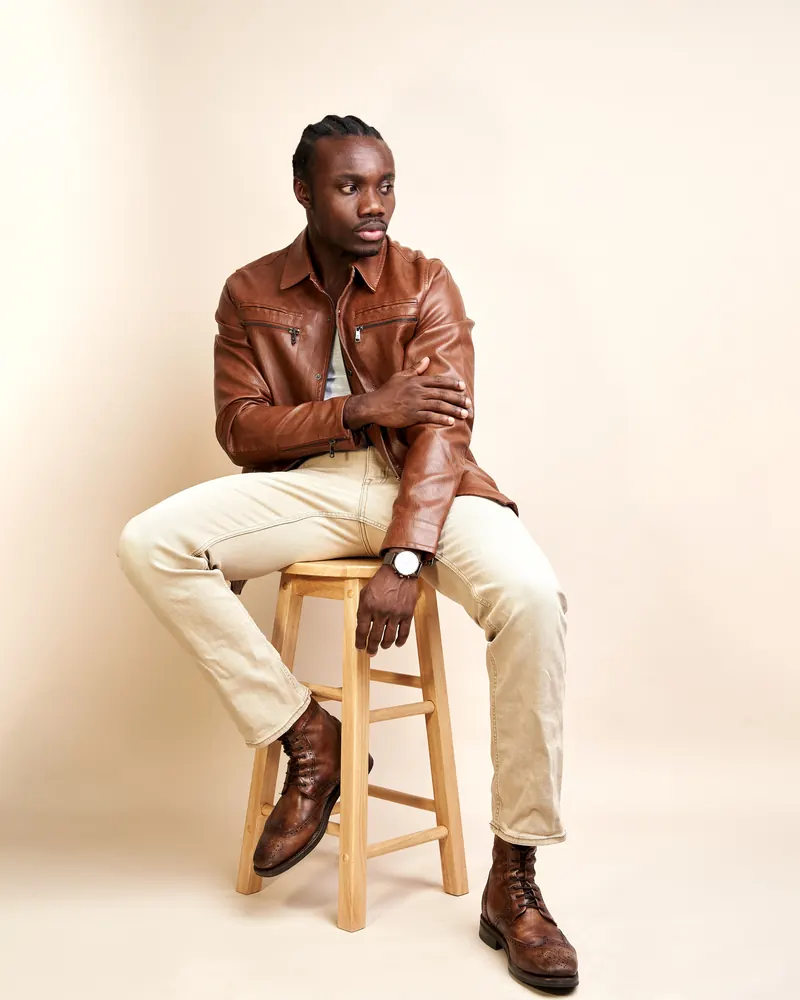 Man in a cognac leather zip-front jacket and stone chinos, seated on a wooden stool against a cream studio backdrop