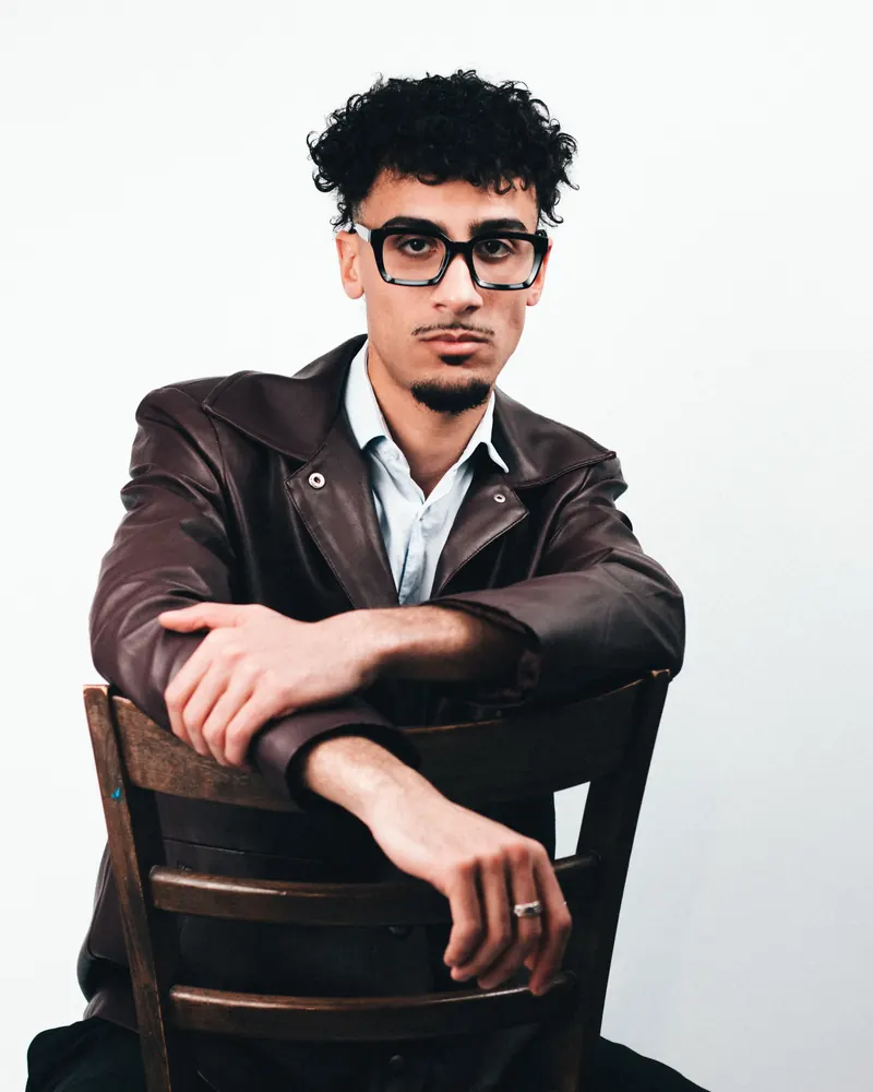 Man in an oxblood leather jacket and pale blue shirt, seated backwards on a wooden chair against a white wall